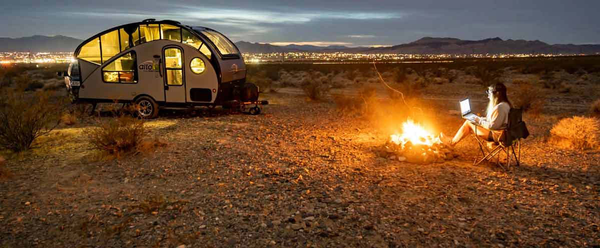 Teardop trailer in the desert with person sitting by blazing campfire with laptop in foreground, and city lights spanning the horizon in background